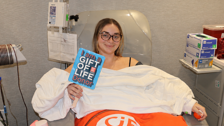 Gift of LIfe stem cell donor Lauren is shown in the collection center on her donation day. She's holding up a small sign that says "I am a Gift of Life donor!" Lauren's younger sister had a bone marrow transplant as a child, so she understood the need for donors and the importance of showing up when she was called as a match.