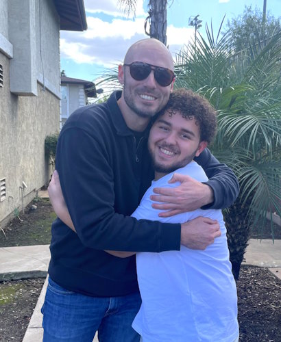 Gift of LIfe stem cell donor Bobby Alexis (left wearing sunglasses) hugs the 18-year-old whose life he saved with a transplant, Aidan Samoon.  Aidan is wearing a white tee shirt, and they are standing outside in front of a palm tree.