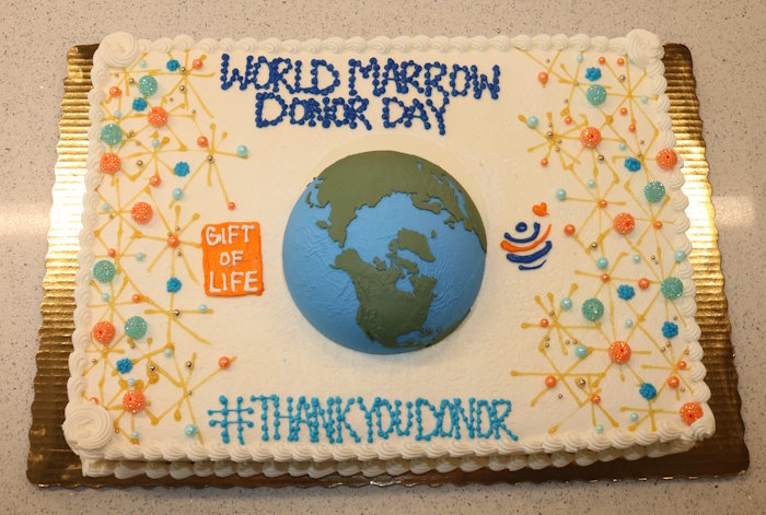 This close-up of the 2026 World Marrow Donor Day Cake at Gift of Life shows a globe in the center, surrounded by white frosted sheet cake sporting the Gift of Life and World Marrow Donor Day logos, plus orange, blue, and gold sparkles.
