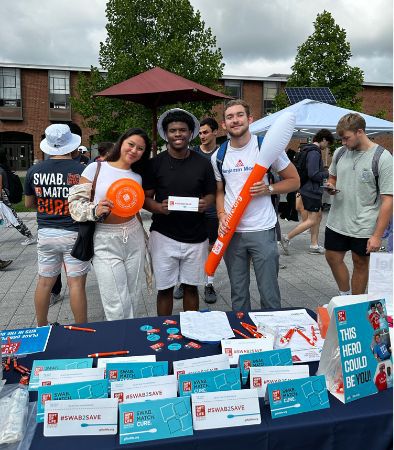 Three female college students who are in the Gift of Life Campus Ambassador Program stand behind their swabbing table, which is laden with swab kits, orange wristbands, pens, and more.