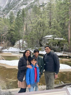 Gift of Life Marrow Registry is seeking a matching stem cell/marrow donor for Shiv Chaudhari, a 46-year-old husband and father whose life is threatened by leukemia. In this photo Shiv, his wife, and two young boys are dressed in winter parkas and scarves, and are at the foot of a snowy mountainside with a stream behind them and pine trees sloping up the hillside. 