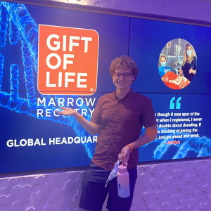 Spencer, a 23-year-old college student from Cal State University - Channel Islands, donated blood stem cells at Gift of Life's collection center to save a man battling leukemia. In this photo he is smiling and standing in front of a bright blue video sign with the orange Gift of Life logo in the entryway to the collection center.