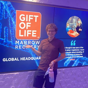Spencer, a 23-year-old college student from Cal State University - Channel Islands, donated blood stem cells at Gift of Life's collection center to save a man battling leukemia. In this photo he is smiling and standing in front of a bright blue video sign with the orange Gift of Life logo in the entryway to the collection center.