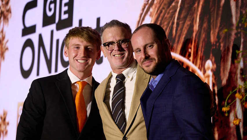 (L-R) Steven Bochco award winner Tommy Hartman, Jesse Bochco, and Sean Flanagan. Tommy is wearing a black suit, a white dress shirt, and an orange necktie. Jesse Bochco wears a camel-colored suite, white dress shirt, and a black tie with gray diagonal stripes. Sean Flanagan Wears a dark blue suit, a medium blue dress shirt and no tie. All are smiling and have their arms around each other. Behind them is a large projection screen showing the Gift of Life logo and a scene of Los Angeles.