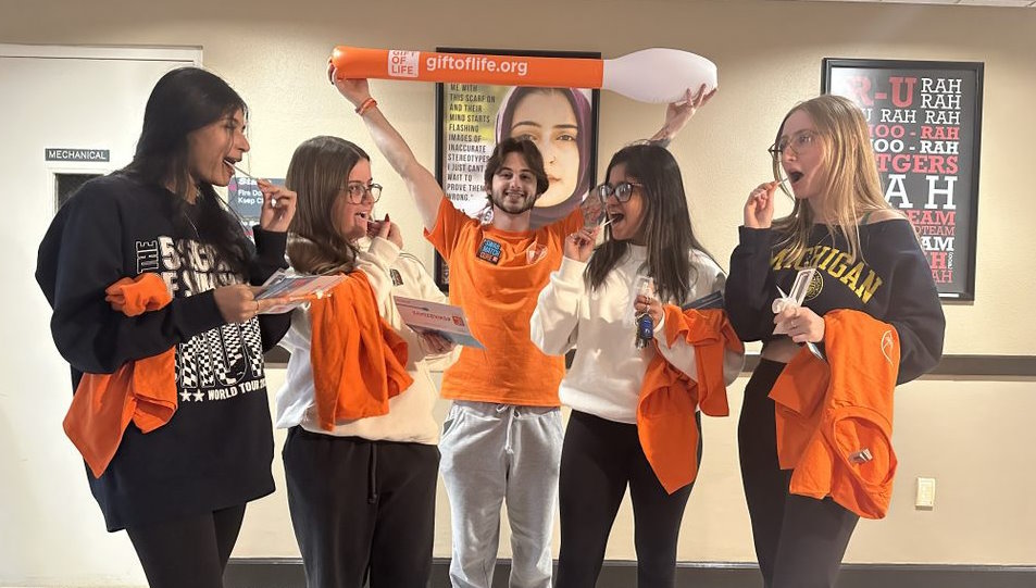 At a Gift of Life Marrow Registry drive a group of female students are using cheek swabs to join the registry while a male student stands behind the group holding an inflatable toy cotton swab over his head. The swab is orange and has the Gift of Life logo on it. 