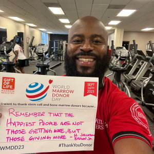 The Clark Atlanta University celebrated World Marrow Donor Day at the PantherFIT gym, and Mr. Marcus Kendrick, a black man in his early 30s, wanted to join in. He is holding up an #UnSelfie sign that reads "Remember that the happiest people are not those getting more, but those giving more," a quote from H. Jackson Brown Jr. 