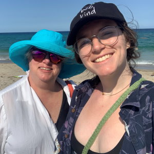 Gift of Life Marrow Registry stem cell donor Sarah and her mom enjoy a day at the beach the day before Sarah donated. Mom is on the left in a turquoise beach hat, and Sarah is on the right wearing a baseball cap. The beach sand and ocean are behind them. Sarah donated her stem cells to save the life of a woman in her 60s battling myelodysplastic disorder, a form of blood cancer. 