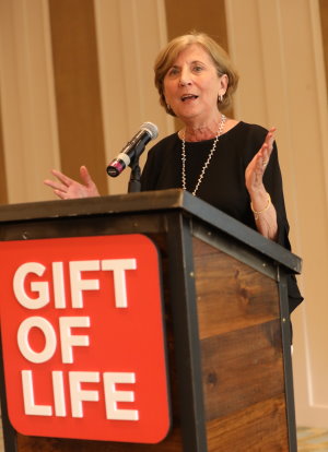 Gail Oliver, a long-time Gift of Life Marrow Registry volunteer, is shown standing at a podium and speaking about her experiences in running recruitment drives for nearly 30 years to help save the lives of blood cancer patients. She is wearing a black dress and has a positive and hopeful expression on her face. 
