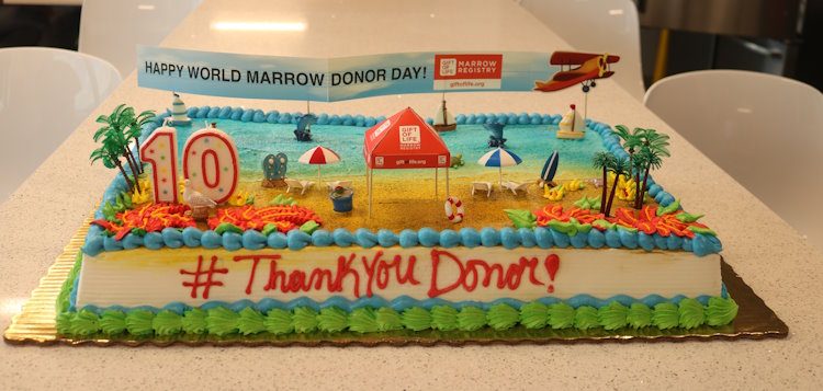 This close up photo of Gift of Life's World Marrow Donor Day cake shows that it is decorated with a beach theme, a large number 10 for the tenth WMDD, a beach tent with the Gift of Life logo, and a tiny plane flying past the beach towing a banner that reads "Happy World Marrow Donor Day"
