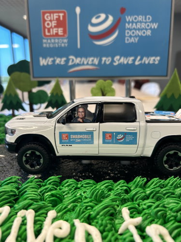 Gift of Life Marrow Registry's entry in the 2023 WMDD cake-off contest features a miniature swabmobile atop the cake, with a tiny Jay Feinberg at the wheel. In the background there is a small blue billboard saying "We're Driven to Save Lives!" and the frosting on the cake is shaped like green grass.  
