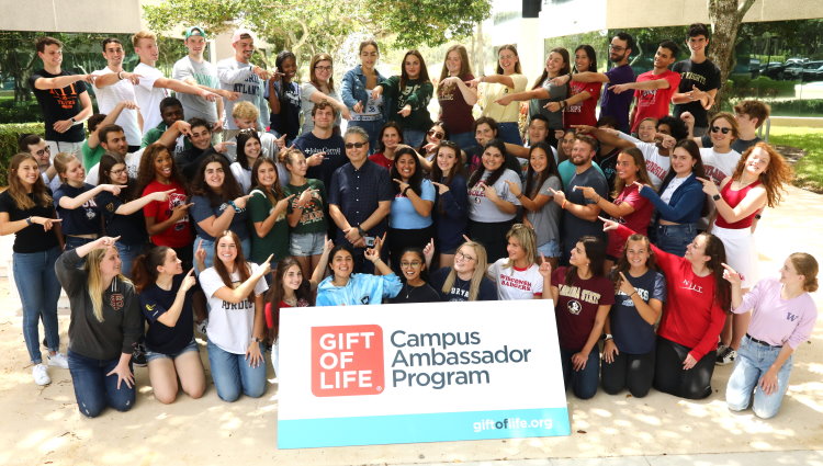 100 Gift of Life participants in the Campus Ambassador Program post with organization CEO Jay Feinberg in front of the headquarters building during the annual training symposium in Boca Raton, Fla. The students are each wearing their alma mater's tee shirt, and all are pointing at Feinberg, who stands in the center of the group smiling. 