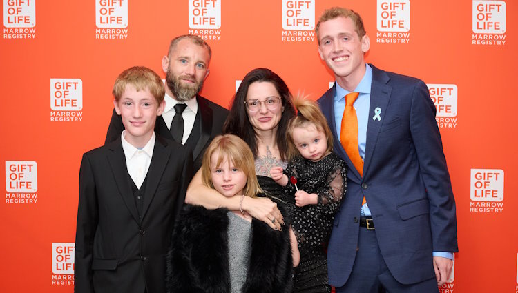 (L-R) Jackson Kobernat, Robert Kobernat, Baelynn Kobernat, Ashley Kobernat, Isla Kobernat (recipient) and Jason Tornes (stem cell donor). The Kobernat family are all dressed nicely in black clothing, and Jason is wearing a blue suit, light blue dress shirt, and orange time. All are smiling and look happy to be meeting each other. Baby Isla has an upright pony tail sticking out of the top of her head, and is holding a red lollypop.  