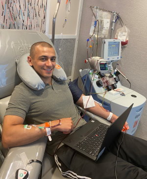 Gift of Life stem cell donor Adam is shown in the organizations collection center as he donates cells for his second matching patient. He has now saved the lives of two people battling to survive leukemia. In this photo he is smiling and working on his laptop computer as he donates stem cells. 