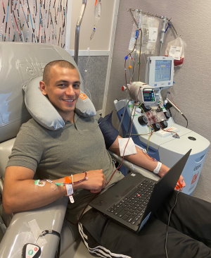 Gift of Life stem cell donor Adam is shown in the organizations collection center as he donates cells for his second matching patient. He has now saved the lives of two people battling to survive leukemia. In this photo he is smiling and working on his laptop computer as he donates stem cells. 