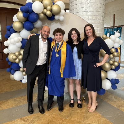 Ignacio, son Lucas, daughter Gabriela, and wife Melissa post in front of an archway of blue, white and gold balloons. Lucas is wearing a royal blue graduation gown with several tassels and ribbons of honor around his neck. 