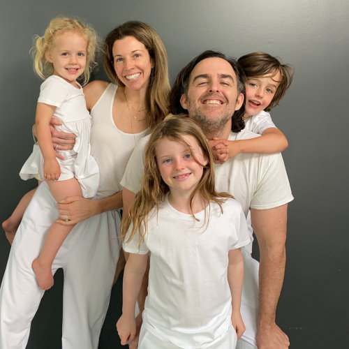 Kirsten Thompson with her husband and children smiling and posing for an informal family portrait. All five are wearing white and are in front of a gray backdrop. 