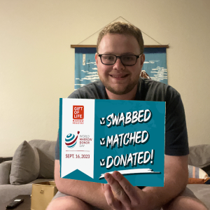Gift of Life Marrow Registry stem cell donor Cole is holding up a "Swabbed, Matched, Donated!" sign for World Marrow Donor Day. He is in his early 20s, has a big smile, sandy hair and a short beard, and glasses. 
