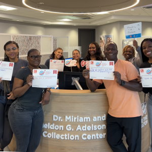 The staff members of Gift of Life Marrow Registry's Adelson Collection Center are gathered around the welcome desk and holding up #UnSelfie signs with their messages of thanks to the stem cell donors who save lives. 