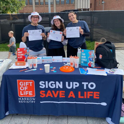 Three Gift of Life Marrow Registry Campus Ambassadors are shown at their recruitment table at Binghamton University as they run a donor drive on World Marrow Donor Day. All three are holding up #UnSelfie signs that are too distant to read. However, all three are smiling and look happy to be there. 