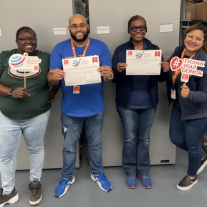 Four of Gift of Life Marrow Registry's logistics staff are posing with #UnSelfie signs with their thanks to marrow and stem cell donors written in. They're relaxed and smiling and look happy. 