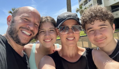 Ignacio, his daugther, wife, and son, outdoors in the sun with their heads close together and big smiles as wife Melissa takes a family selfie of them. 