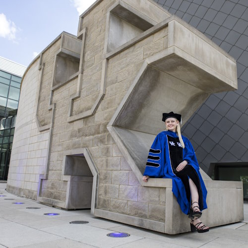 Gift of LIfe stem cell donor Mary is shown in her blue and black doctoral student cap and gown, sitting on the giant cement letters  UK for the University of Kentucky. The famous letters sit outside the Gatton Student Center. Mary succesfully defended her Ph.D. thesis just weeks after donating peripheral blood stem cells to save the life of a man in his early 20s battling leukemia.