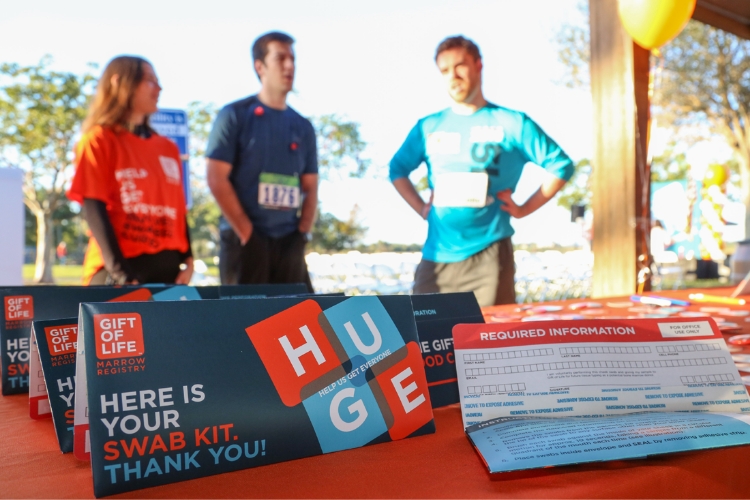 A close-up photo of a recruitment table with many Gift of Life swab kits in the foreground, while three volunteers in their early 20s stand in the background wearing Gift of Life shirts.