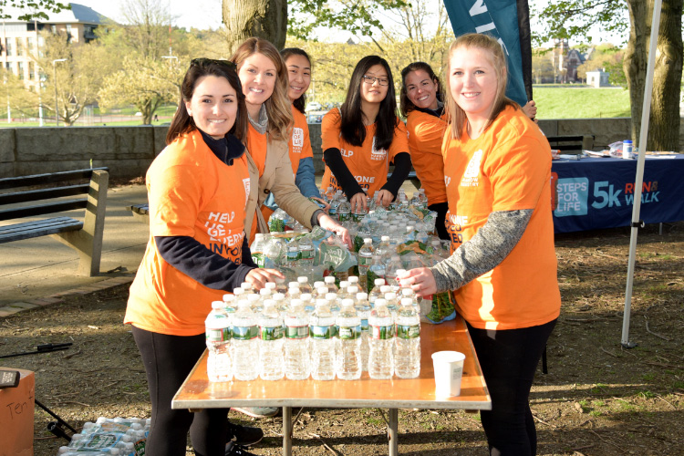 A group of six young women volunteers wearing orange Gift of Life shirts set up water bottles on a table at one of the organization's 5k run or walk fundraising events. Call our main phone number and ask for the Volunteer Coordinator to become involved.