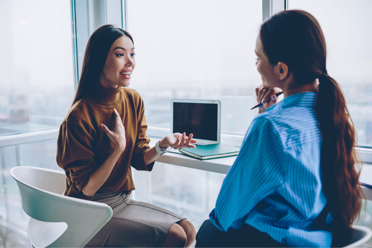 Two young women sit at a desk talking, one is explaining something while the other holds a pen and takes notes. This illustrates the Donor Information Session that Gift of Life has with every donor when they are called as a match for a patient.