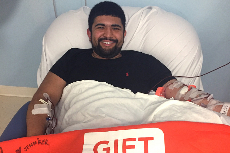 A smiling young Hispanic man sits in a donation chair giving stem cells for his matching recipient. Blood is being drawn from his left arm, the stem cells are being collected by the apheresis machine, and his remaining blood is being returned via his right arm.